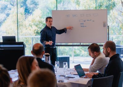 David Jenyns presenting business systems workshop to seated attendees at a whiteboard in a modern glass-walled room