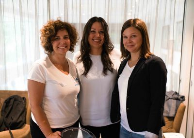 Three women smiling together at a SYSTEMology event, two wearing branded white SYSTEMology t-shirts