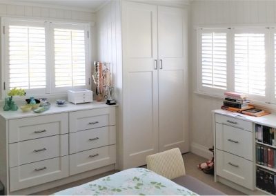 White built-in wardrobe with recess panel doors, chest of drawers, and plantation shutters in a bright bedroom