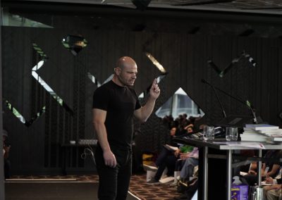 Male speaker in black t-shirt gesturing with one finger raised at a business summit event with audience seated behind