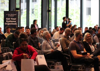 Business owners attending a SYSTEMology summit seated in a conference room with a 'Get Your Systems Organised' banner